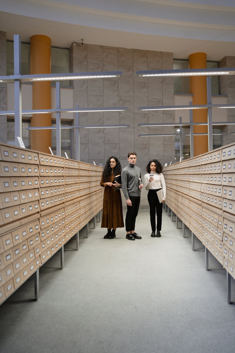 Three students standing in a library card catalog room, exploring resources for research.