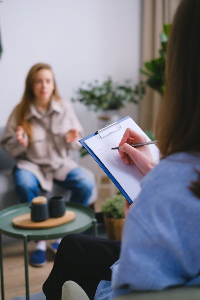 Unrecognizable female psychologist taking notes on clipboard while listening to patient sitting on blurred background during psychotherapy consultation in organisation